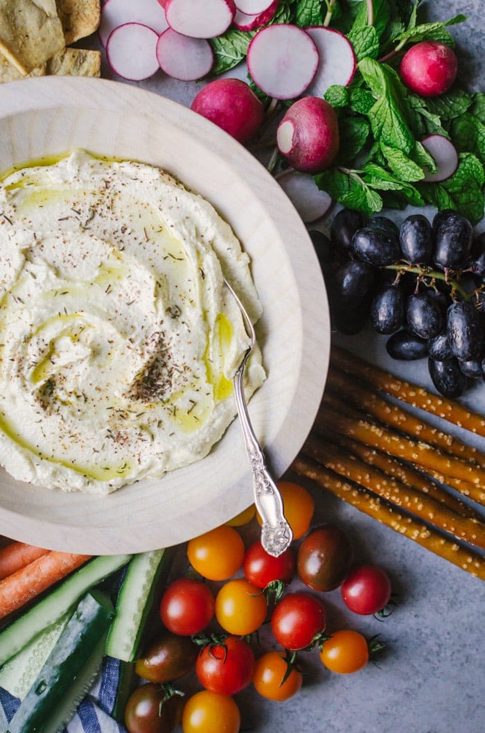 Dip in wooden bowl with spoon surrounded by fresh fruits and vegetables