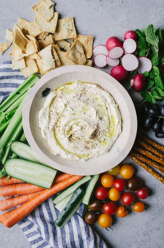 Dip in bowl next to linen and surrounded by fruits, vegetables, and pita chips