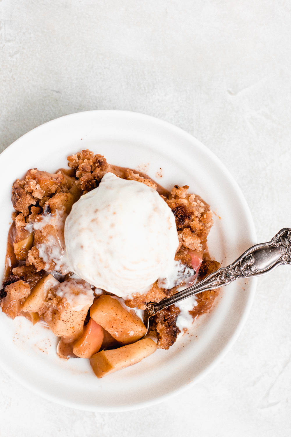 apple crumble in white bowl with spoon topped with vanilla ice cream