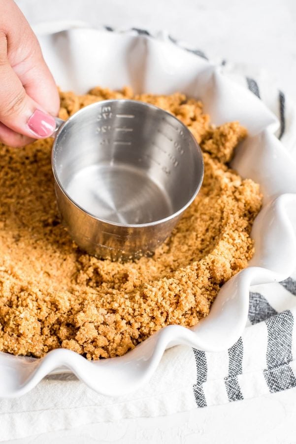 graham cracker crumbs being pressed into pie pan with back of measuring cup