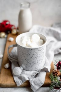 white enamel mug on wood board near Christmas decorations filled with white hot chocoalte and marshmallows